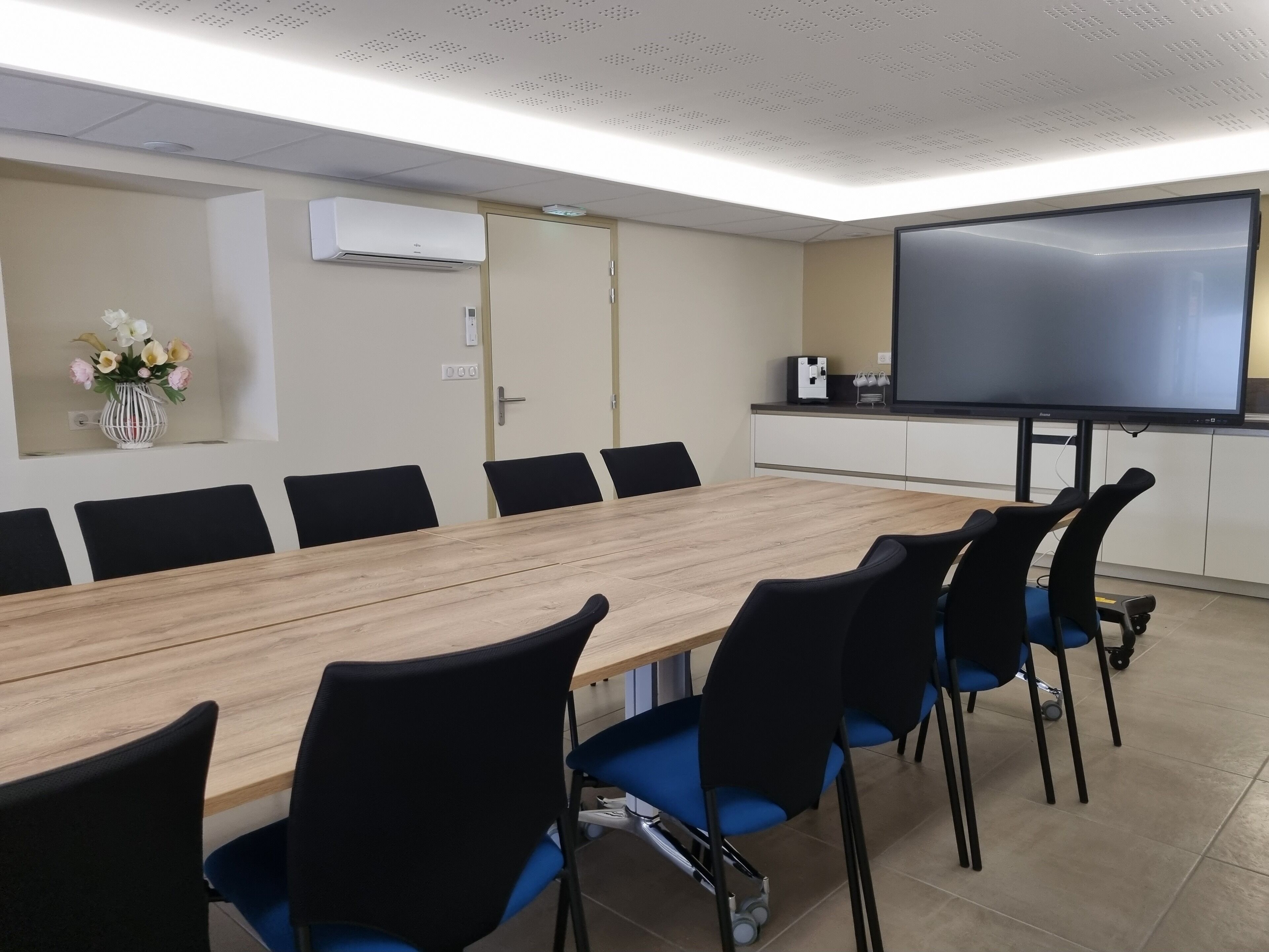Ein Konferenzraum mit einem langen Holztisch, schwarzen Stühlen, einem großen Flachbildfernseher und einer weißen Klimaanlage an der Wand.