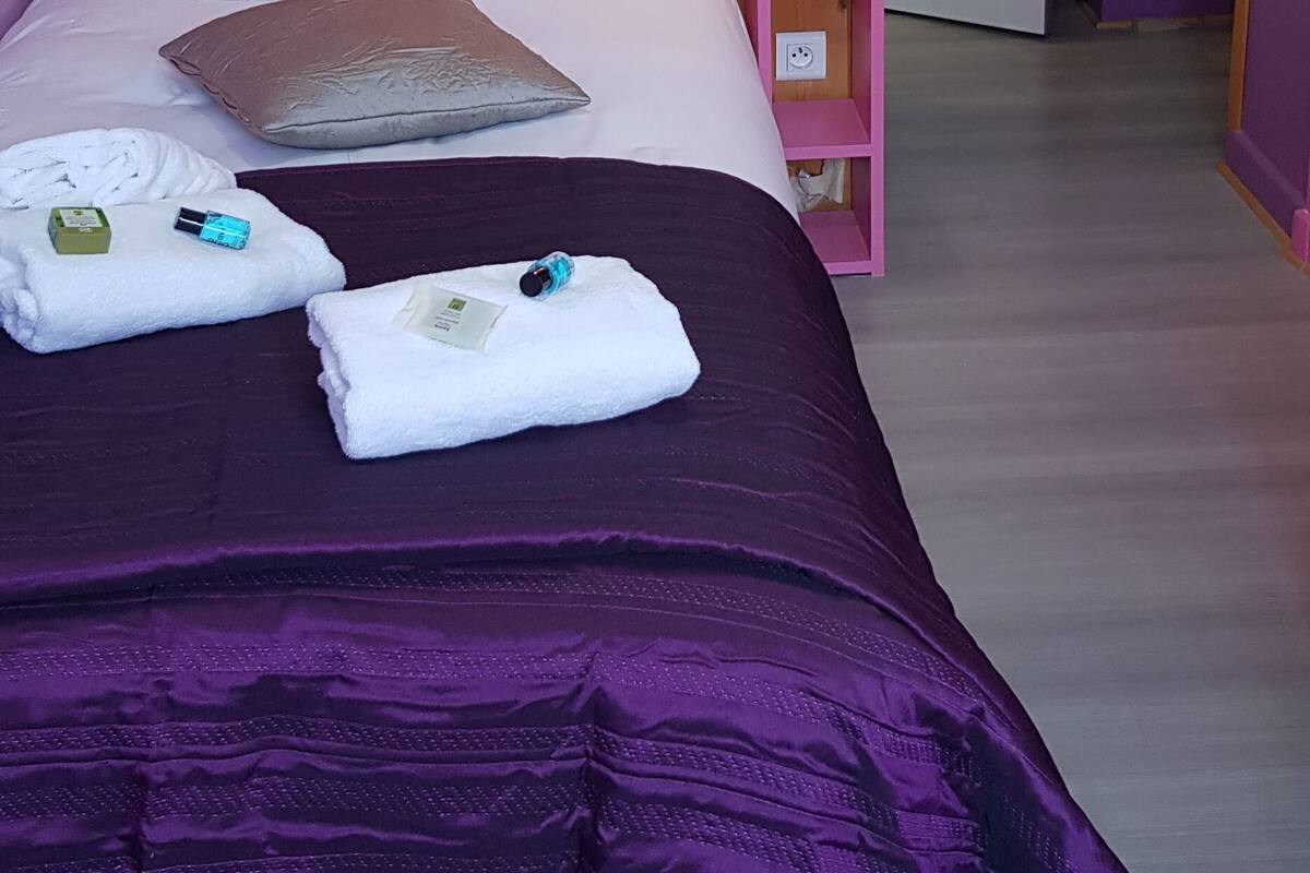 Une chambre avec une tête de lit en bois, un mur rose et une couvre-lit violette. On y trouve des serviettes et une petite table de chevet rose avec une lampe.