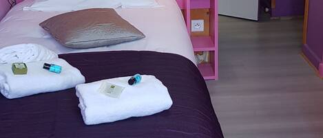 A room with a wooden headboard, a pink wall, and a purple bedspread. There are towels and a small pink nightstand with a lamp.
