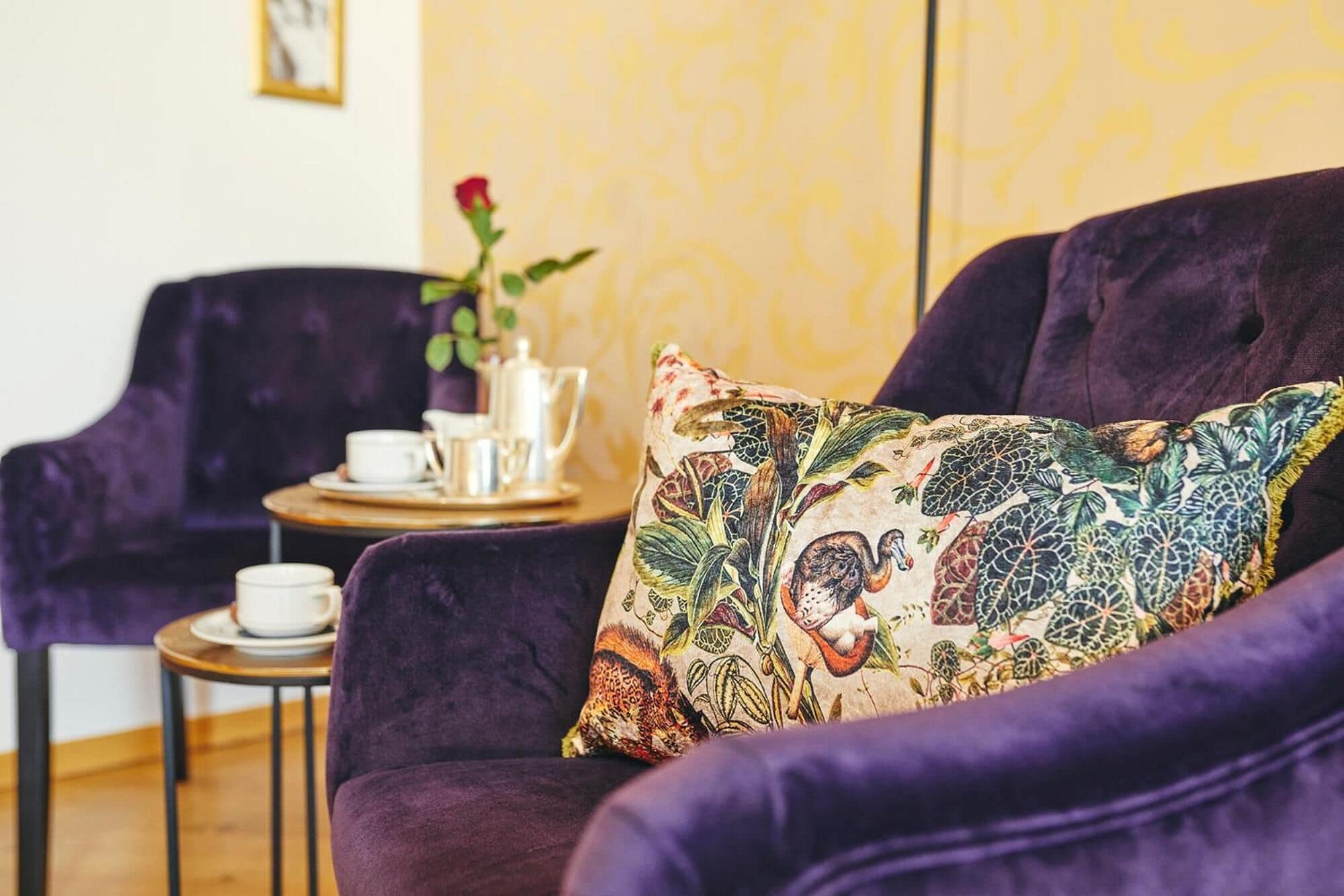 Un sofá púrpura con un cojín floral de colores, una mesita redonda con una taza y platillo blancos, y un florero con una sola rosa roja.