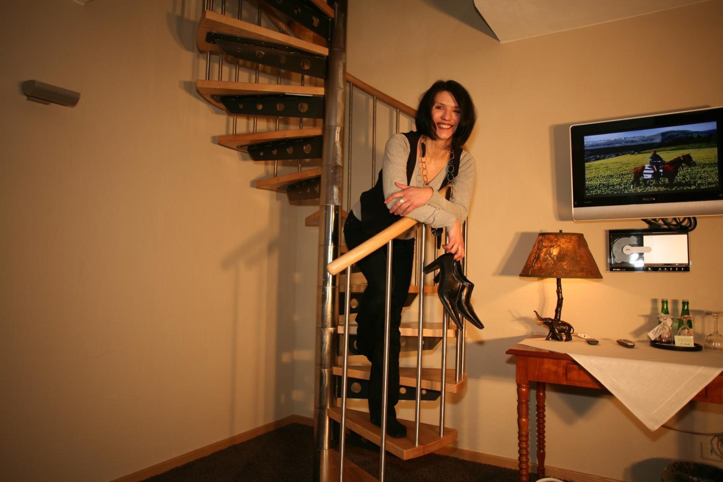 A person standing on a spiral staircase in a room with a television displaying a horse riding scene, a lamp, and a small table with bottles.