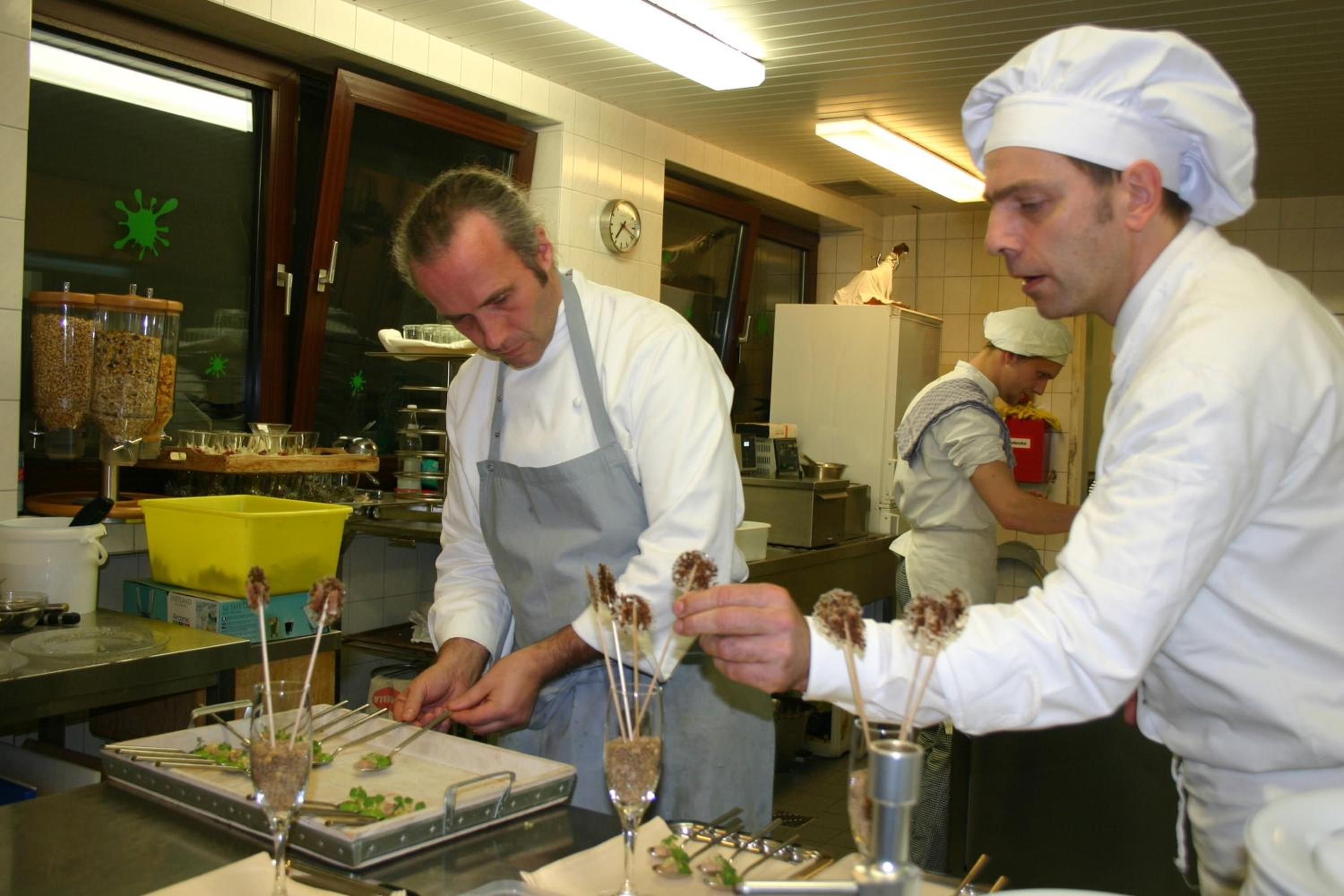 Chefs preparando comida en una cocina con mostradores de acero inoxidable y diversos utensilios de cocina.