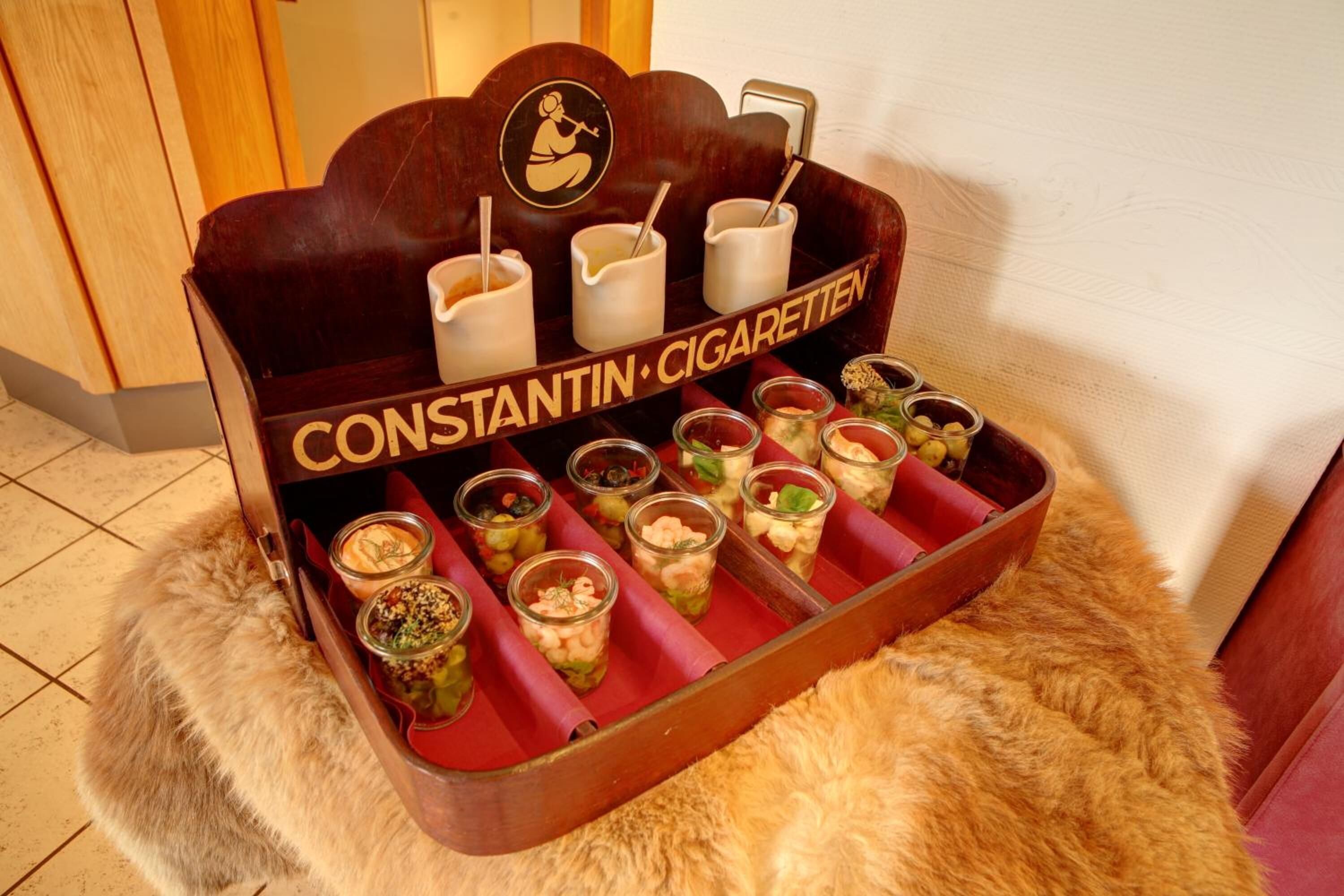 A wooden tray with multiple compartments containing various food items and a sign that reads "CONSTANTIN CIGARETTEN".