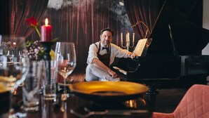 Un homme joue du piano à queue dans une pièce faiblement éclairée, avec une bougie sur la table et une rose sur le côté gauche.