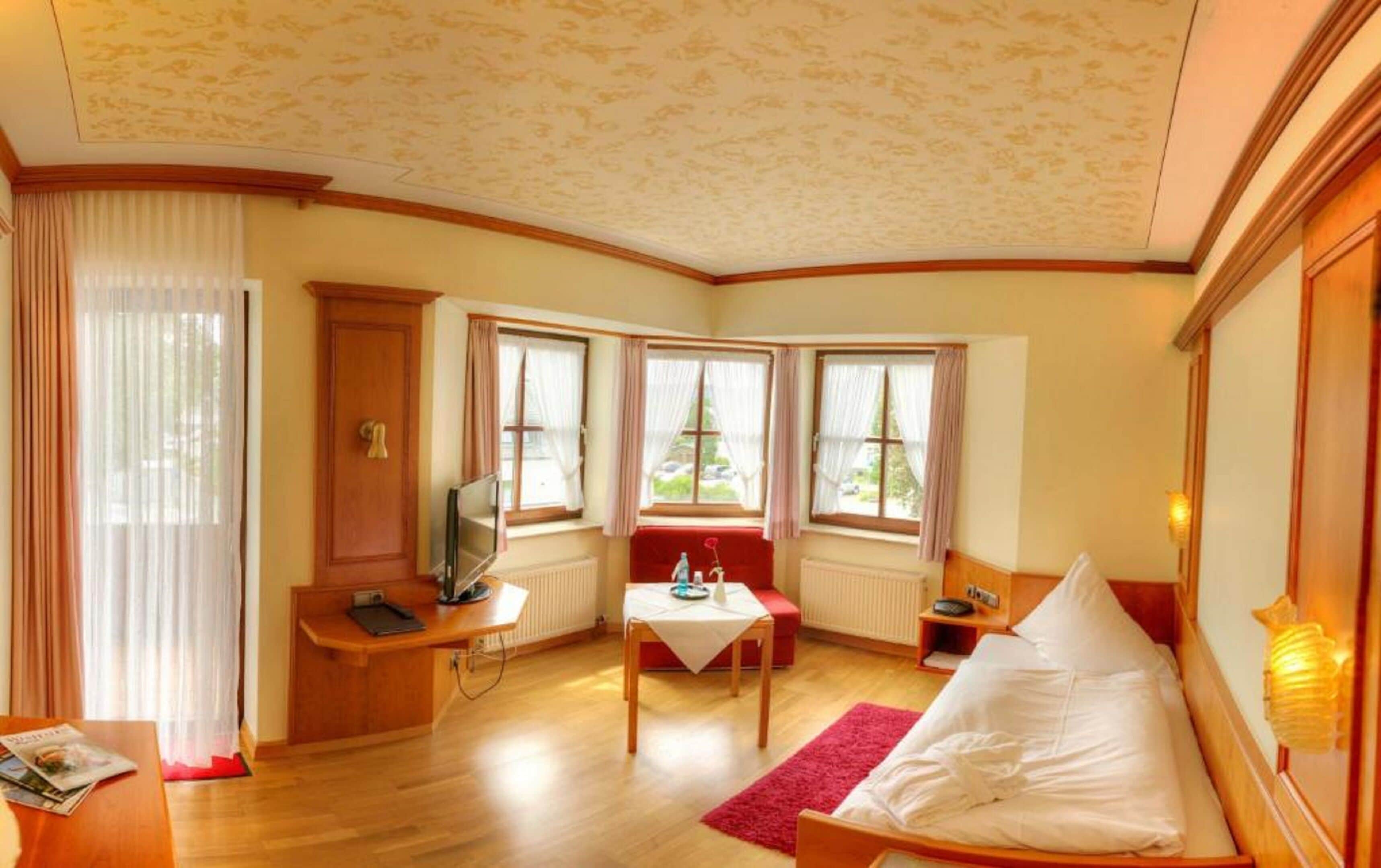 A hotel room with a wooden desk, a television, a red bench, and a bed.