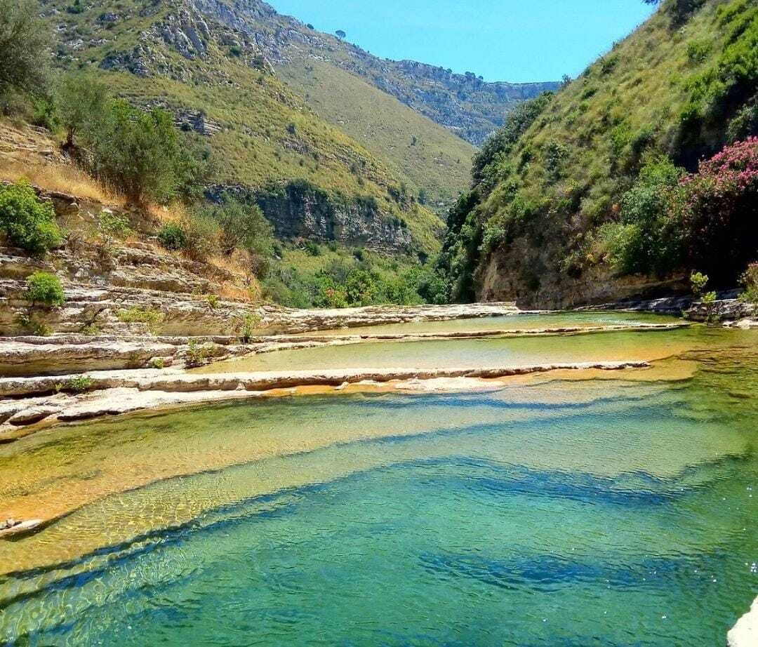 A clear, natural pool surrounded by rocky terrain and greenery.