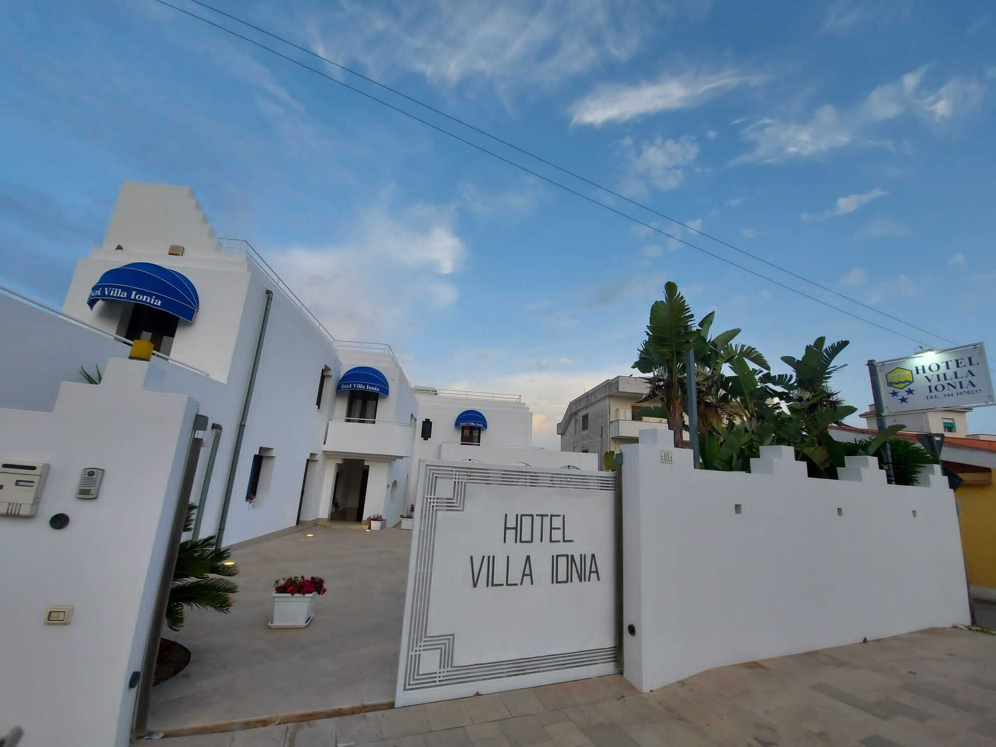 A white building with a sign reading "Hotel Villa Ionia" and a blue awning.