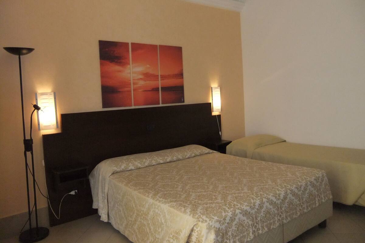 Hotellihuone, jossa on suuri sänky, kaksi yöpöytää, lattiavalaisin ja seinälle ripustettu taideteos.