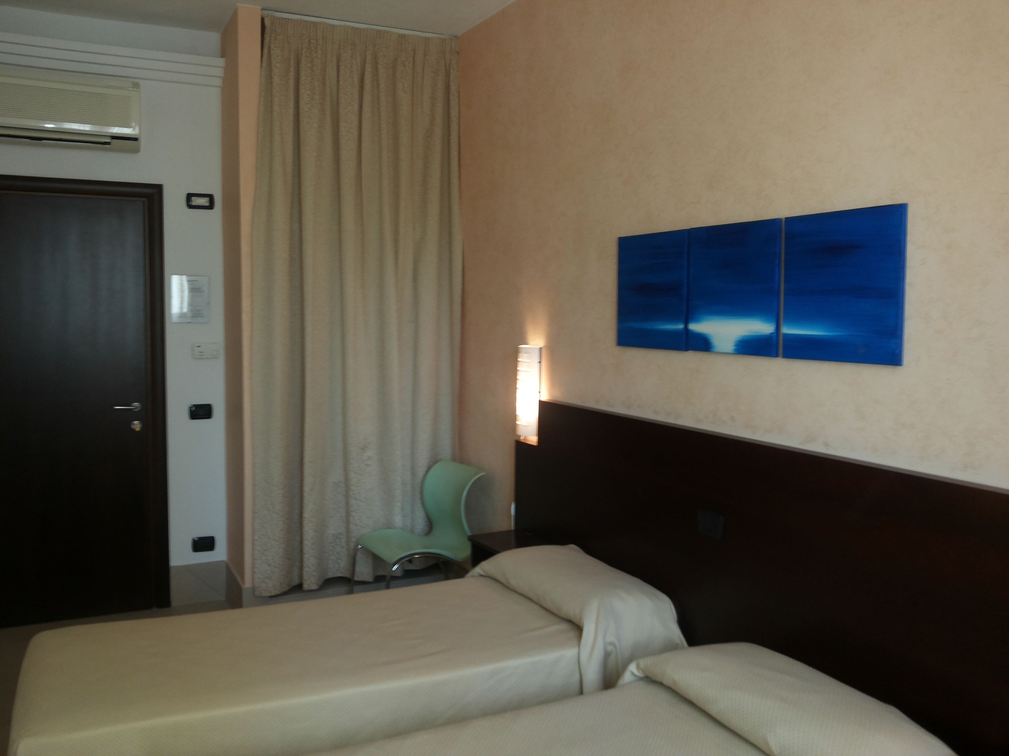 A hotel room with two beds, a window with curtains, a wall-mounted light, and a painting.