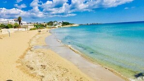 A beach with clear turquoise waters, sandy shore, and buildings in the background. - Hotel Villa Ionia (Avola)