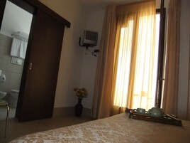 Una habitación de hotel con una cama, un televisor montado en la pared, un florero con flores y una ventana con cortinas.
