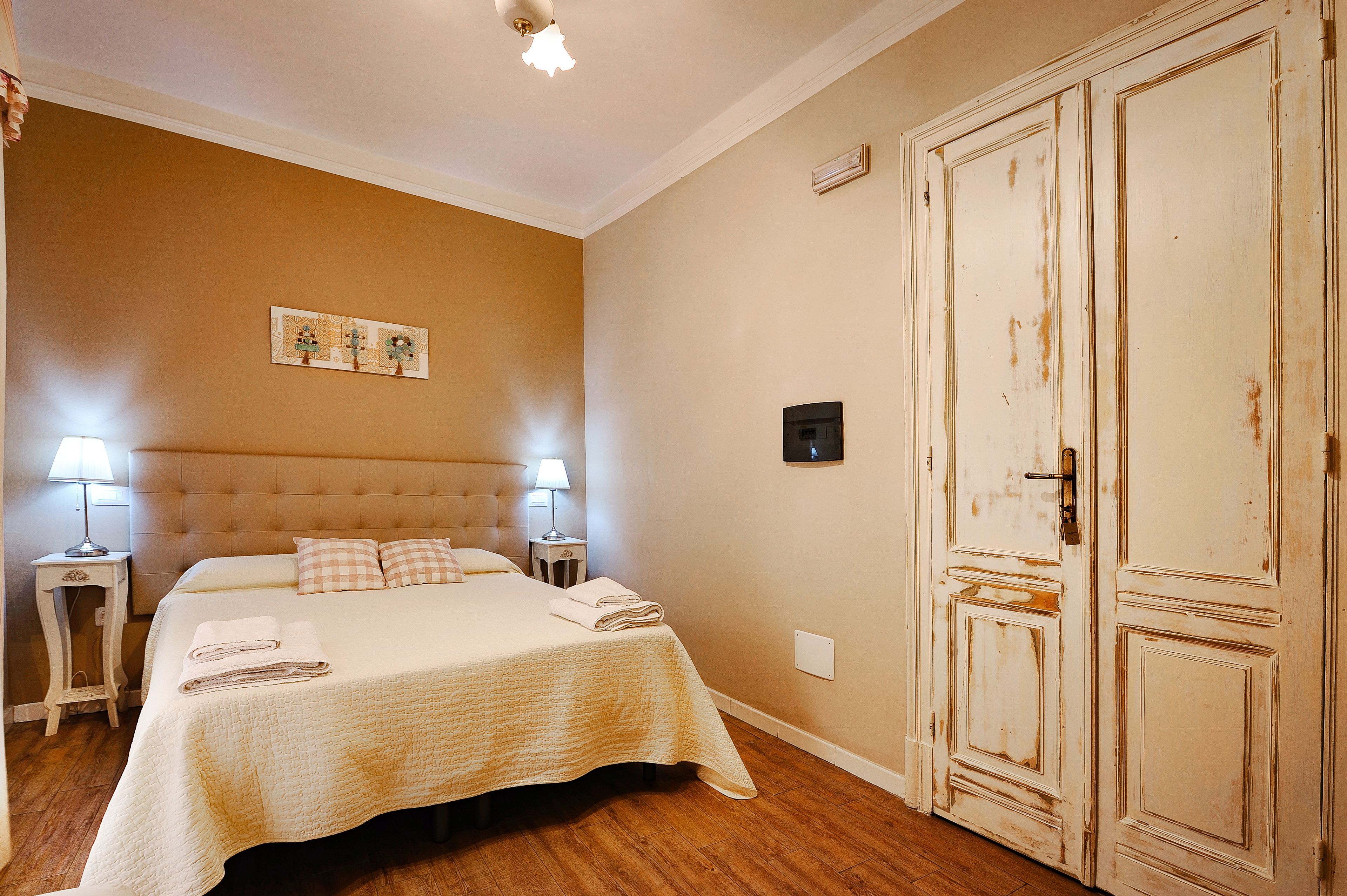 A hotel room with a large bed, two bedside tables with lamps, a wardrobe, and a wall-mounted air conditioning unit.