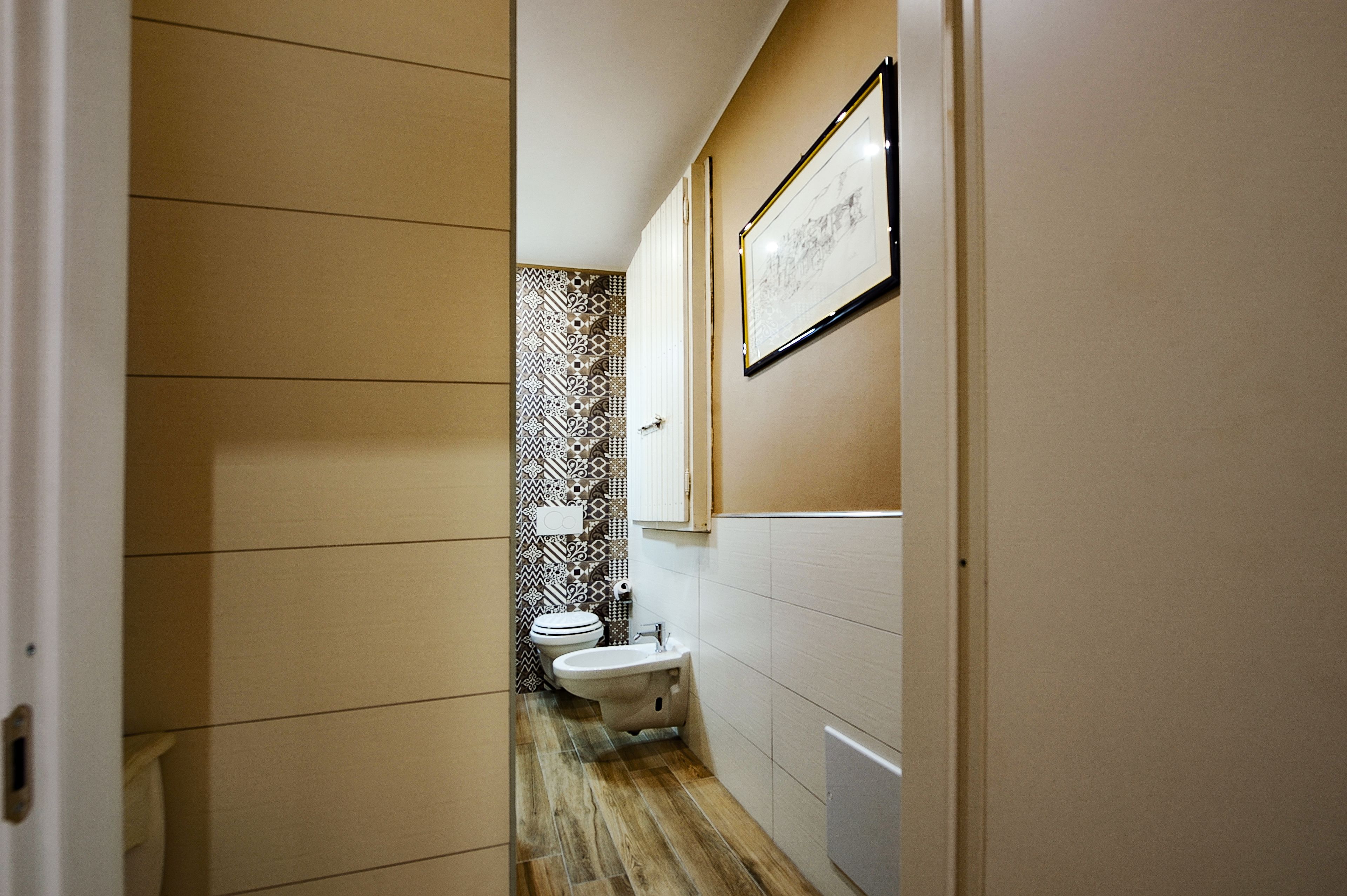 Un couloir étroit avec des toilettes et un bidet, un tableau encadré au mur et des carreaux muraux à motifs.