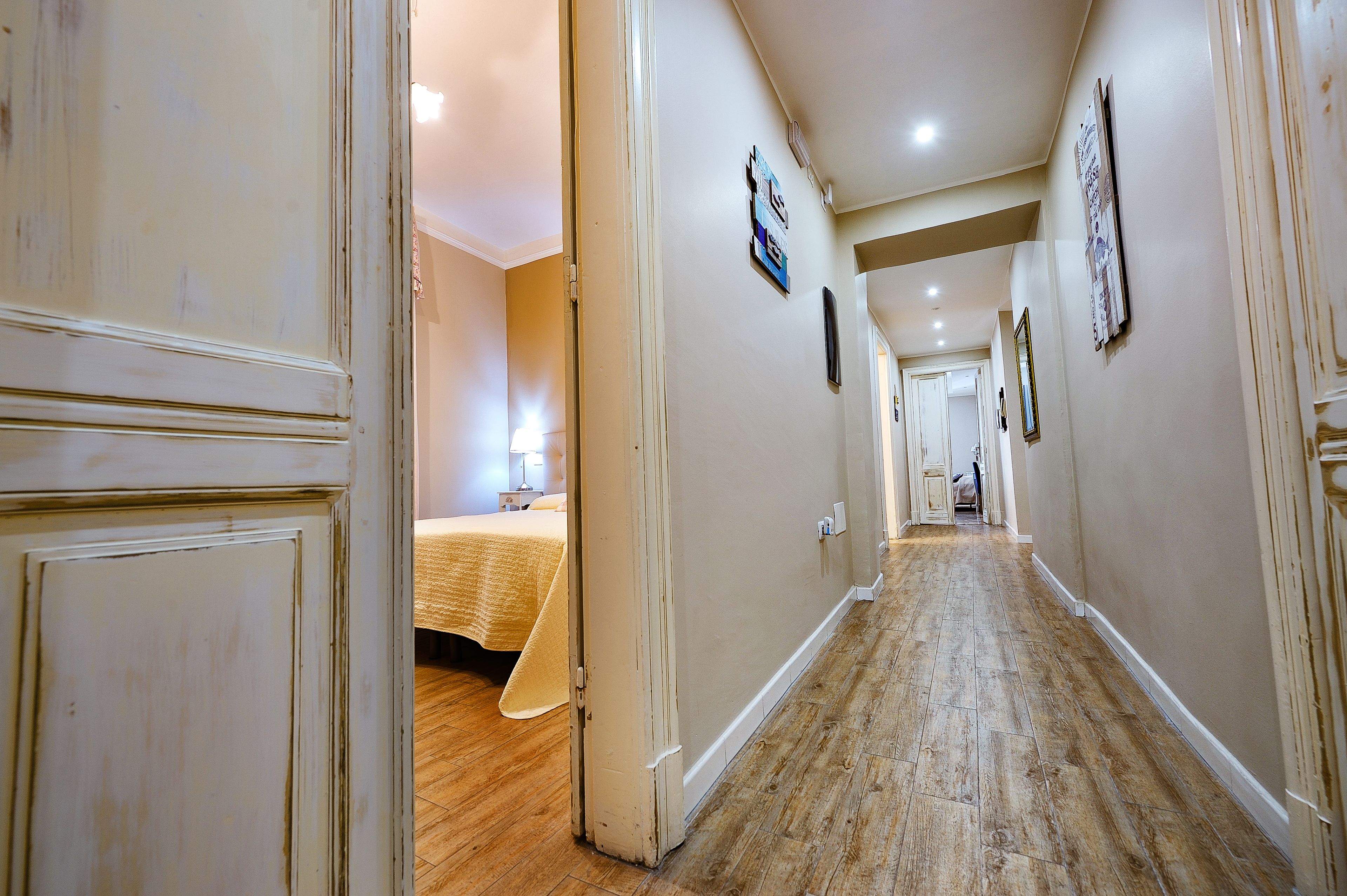 A hotel hallway with wooden flooring, cream walls, and a bed with a yellow bedspread in the background.