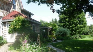 Une maison en pierre avec un toit recouvert de tuiles rouges, un chemin de pierre menant à l’entrée et un jardin luxuriant où poussent diverses plantes et fleurs.