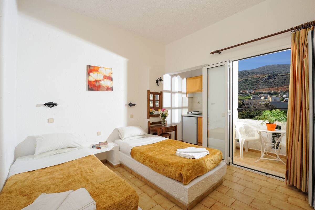 A hotel room with two beds, a desk, a chair, and a balcony with a view of the countryside.
