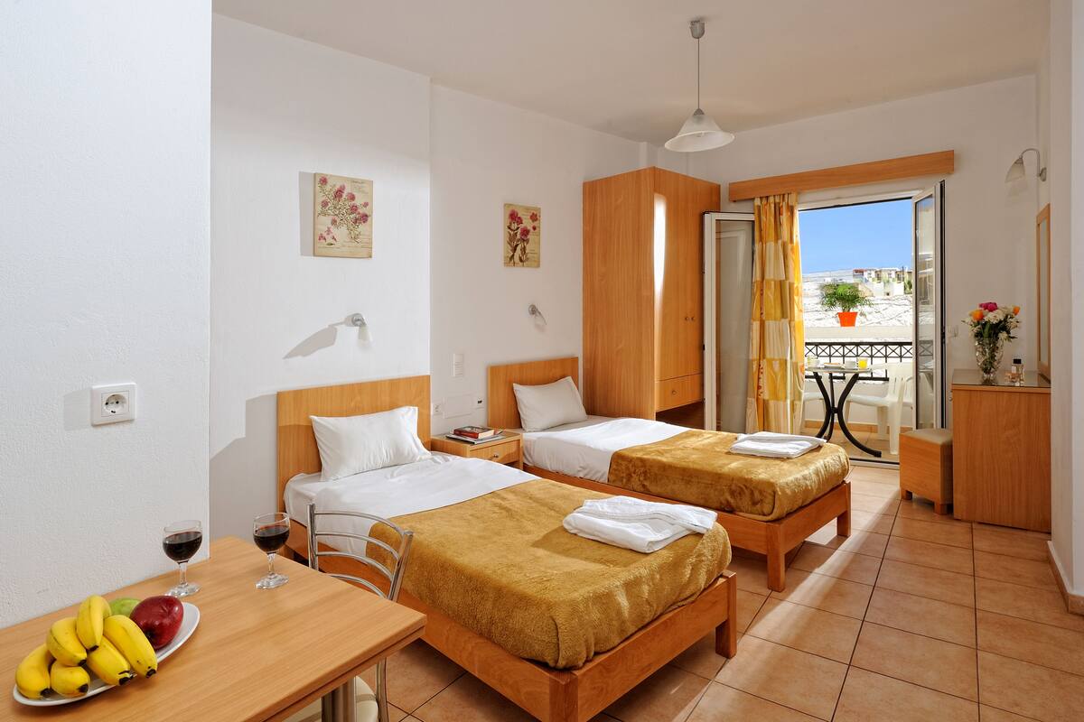 A hotel room with two beds, a dining area with a table and chairs, a small balcony with a table and chairs, and a view of the outside.