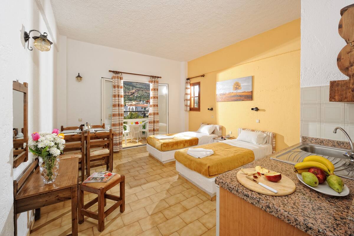 A compact hotel room with two beds, a dining area, a kitchen with fruit, and a balcony with a view.