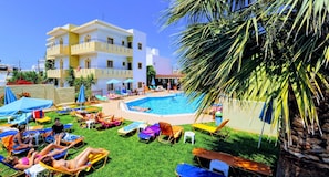 Un espace aménagé au bord de la piscine, avec plusieurs chaises longues et des parasols, une piscine, et un immeuble à plusieurs étages en arrière-plan.