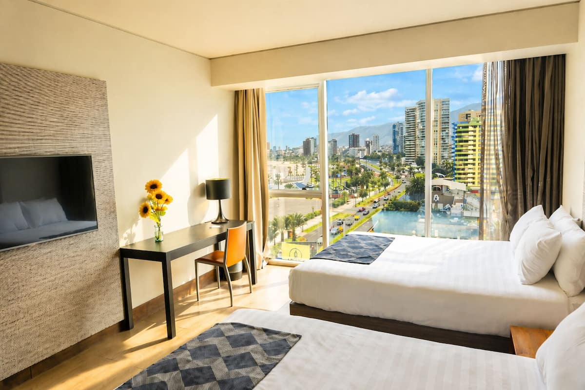 A hotel room with two beds, a desk, a chair, a TV, and a large window with a city view.