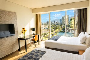 Ein Hotelzimmer mit zwei Betten, einem Schreibtisch, einem Stuhl, einem Fernseher und einem großen Fenster mit Blick auf die Stadt.