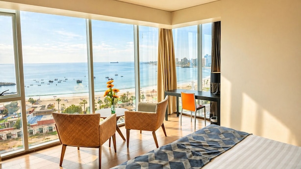 Quarto de hotel com uma grande janela, uma cama, duas cadeiras, uma mesinha com um vaso de flores e uma escrivaninha com abajur.