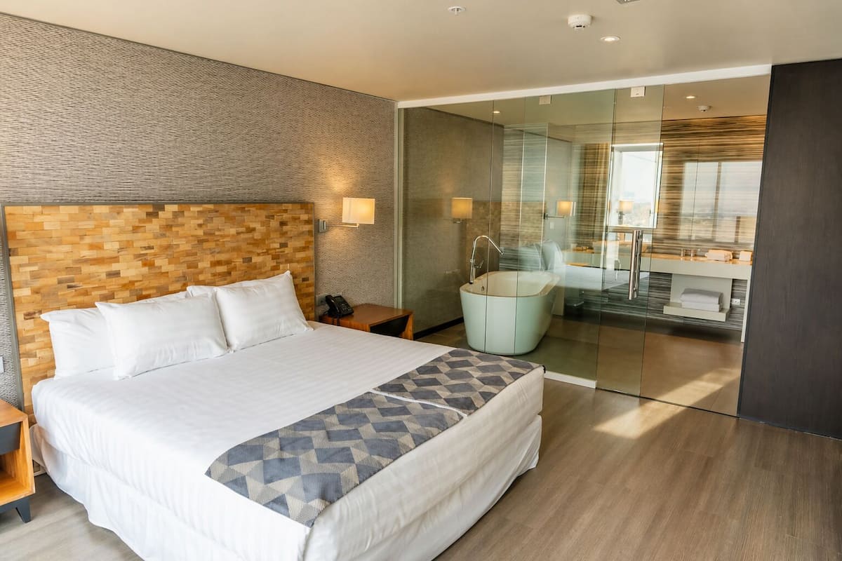 A modern hotel room with a large bed, a glass-enclosed bathroom, and a wooden headboard.