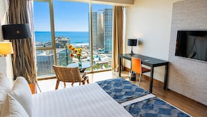 Quarto de hotel com cama, escrivaninha, cadeira, televisão e uma grande janela com vista para a cidade.