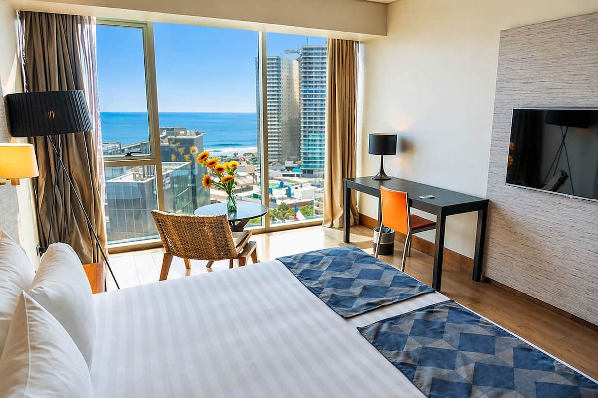 A hotel room with a bed, a desk, a chair, a TV, and a large window with a city view.