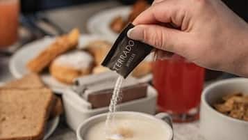 Una mà escampant pols en una tassa de cafè, amb una plat de torrades i un got de beguda vermella al fons.