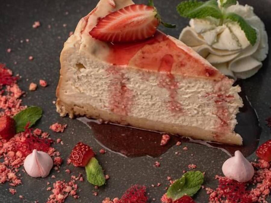 A slice of cheesecake with strawberry and mint garnish on a dark plate.