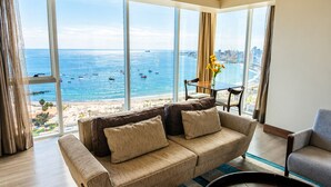 Uma sala de estar moderna com uma grande janela que dá para a praia e o horizonte urbano, um sofá, uma poltrona e uma mesinha com um vaso de flores.