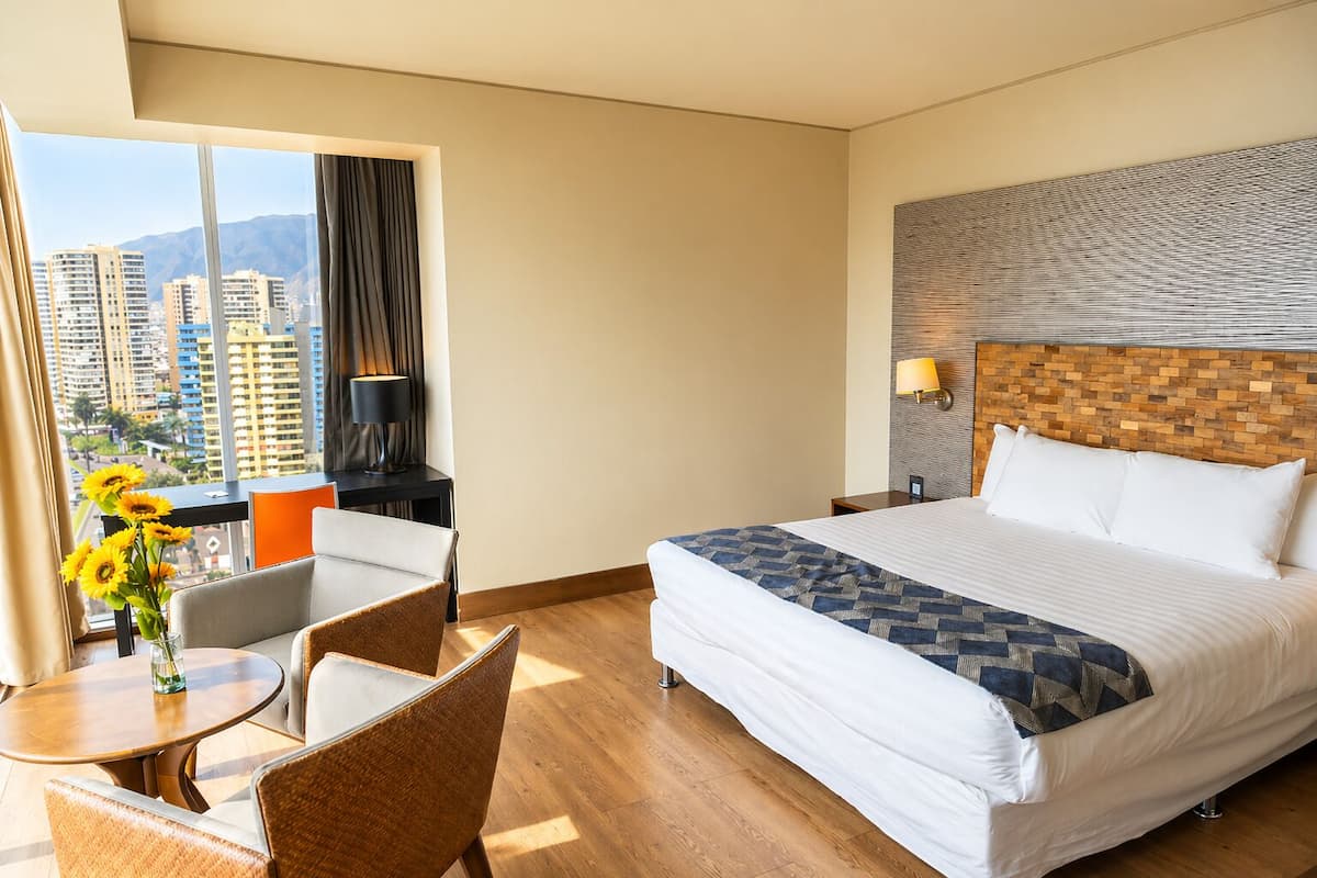 A hotel room with a large bed, a desk with a chair, a view of high-rise buildings, and a decorative wall.