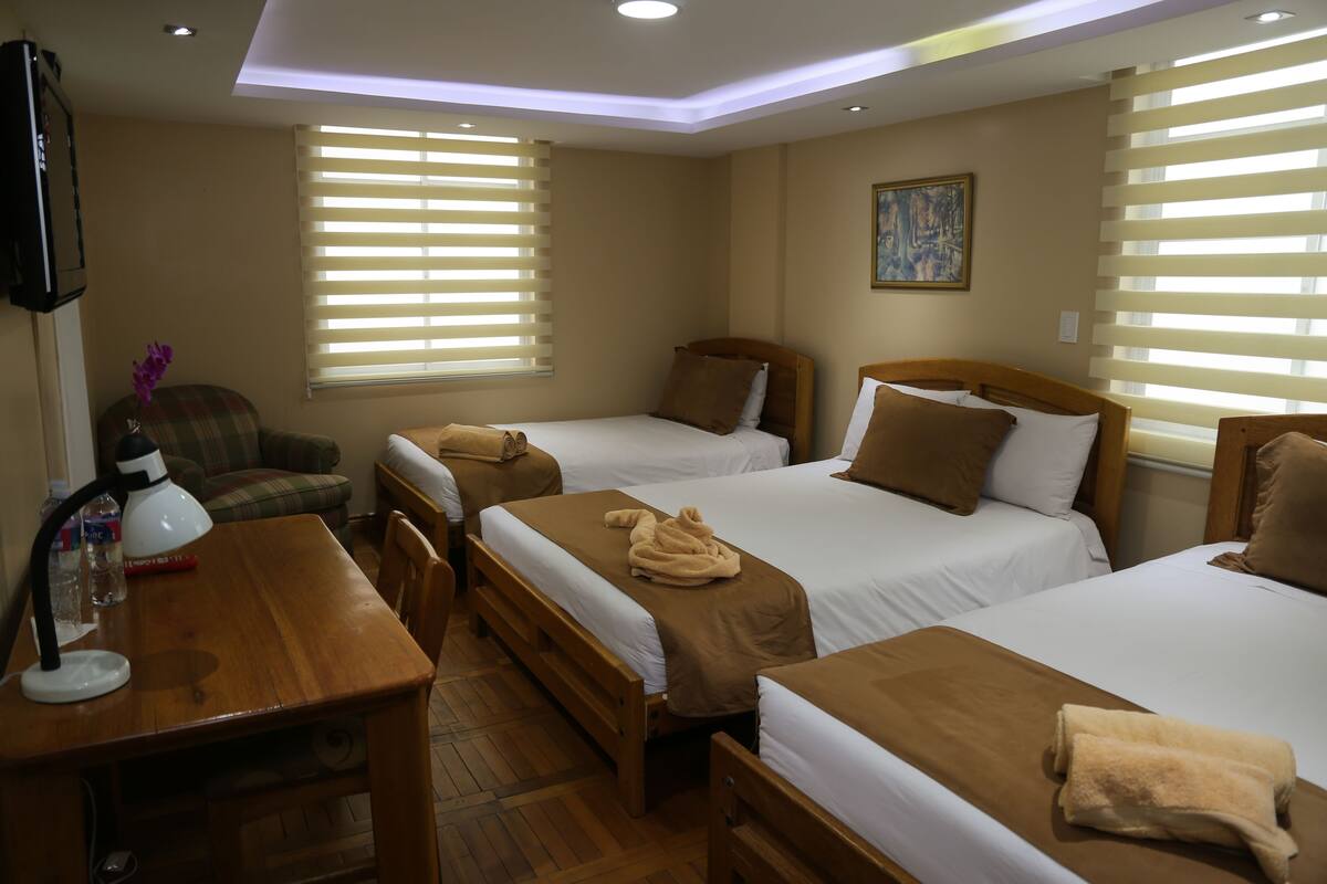 A hotel room with three beds, a desk with a lamp, a chair, and a painting on the wall.