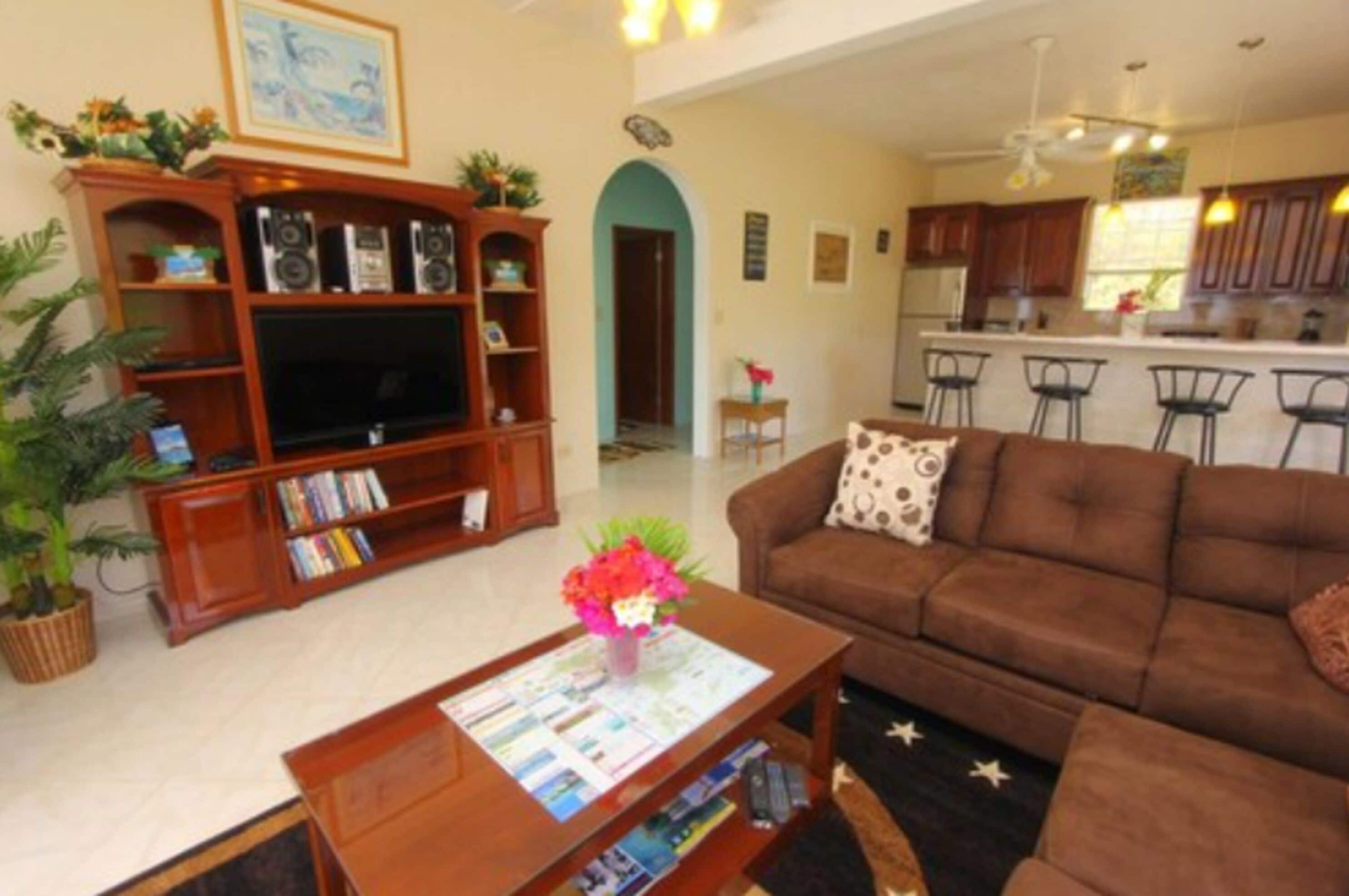 A living room with a brown sofa, a wooden entertainment unit with a TV, and a coffee table with magazines. There is a kitchen visible in the background with bar stools.