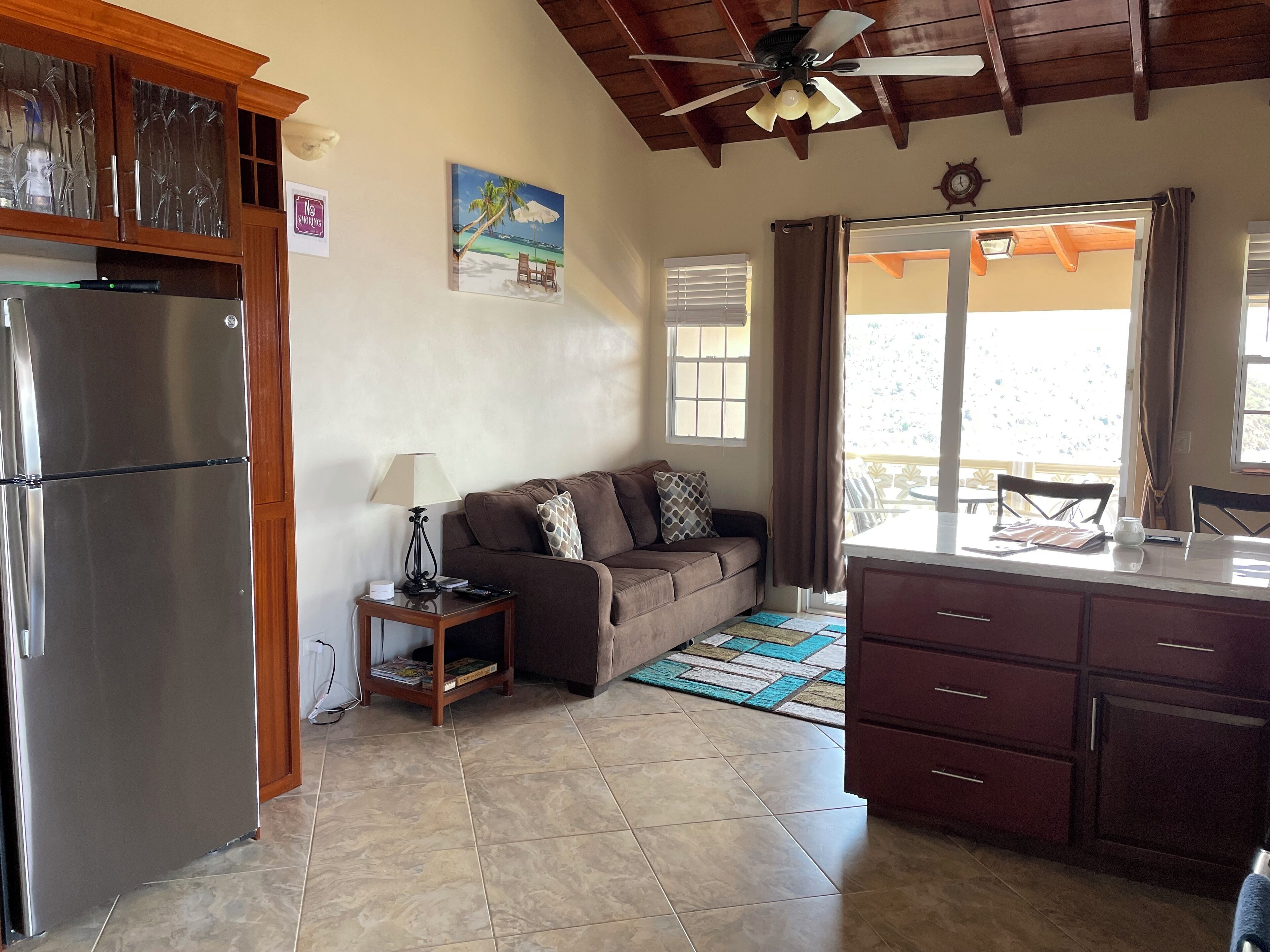 Cocina compacta con refrigerador, gabinetes de madera, zona de comedor con mesa y sillas, sofá y ventilador de techo.