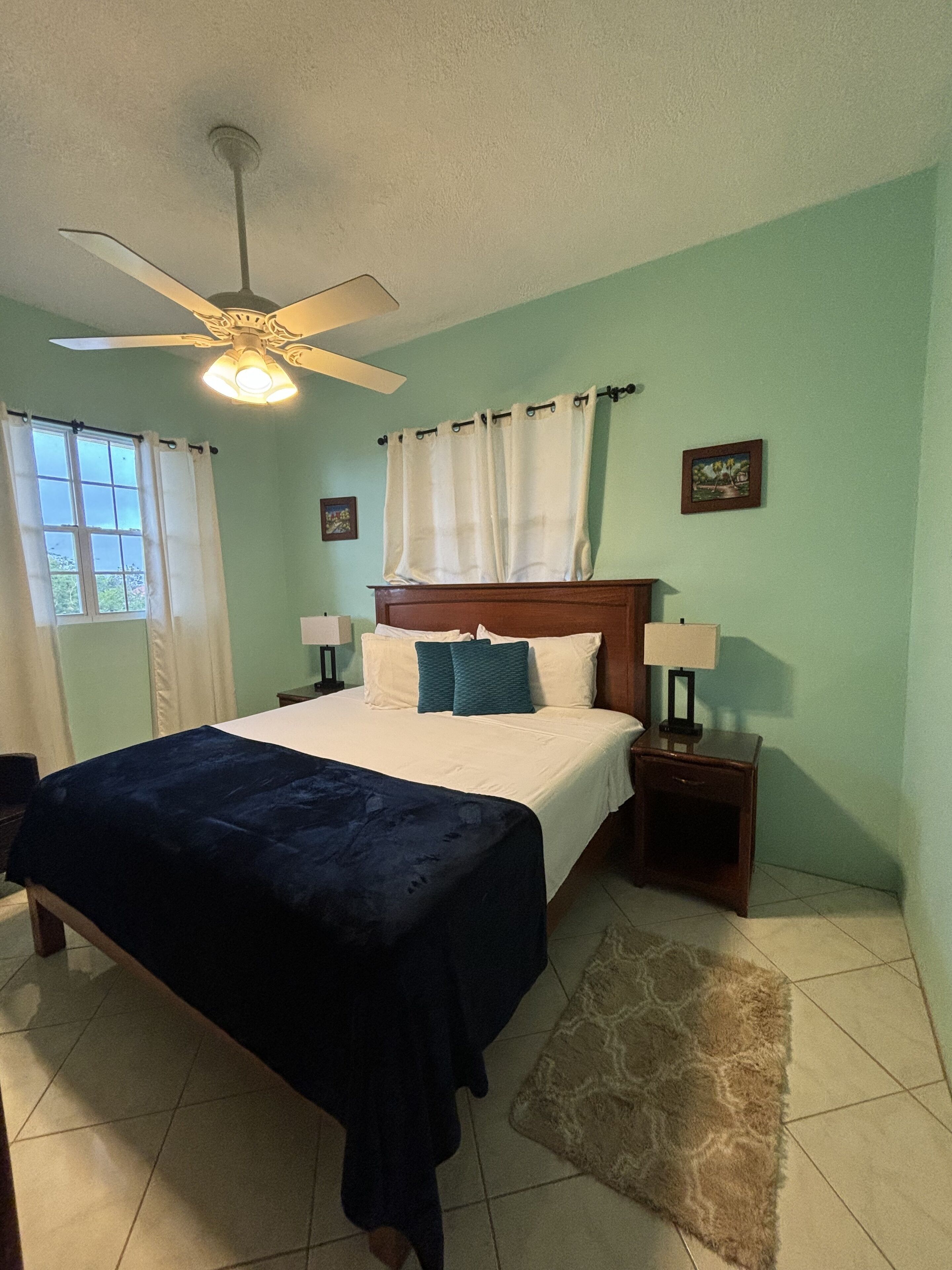 A bedroom with a bed, two bedside tables, a ceiling fan, a window with curtains, and a rug.
