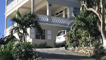 Un edificio de dos plantas con balcón, entrada para coches y vegetación alrededor.