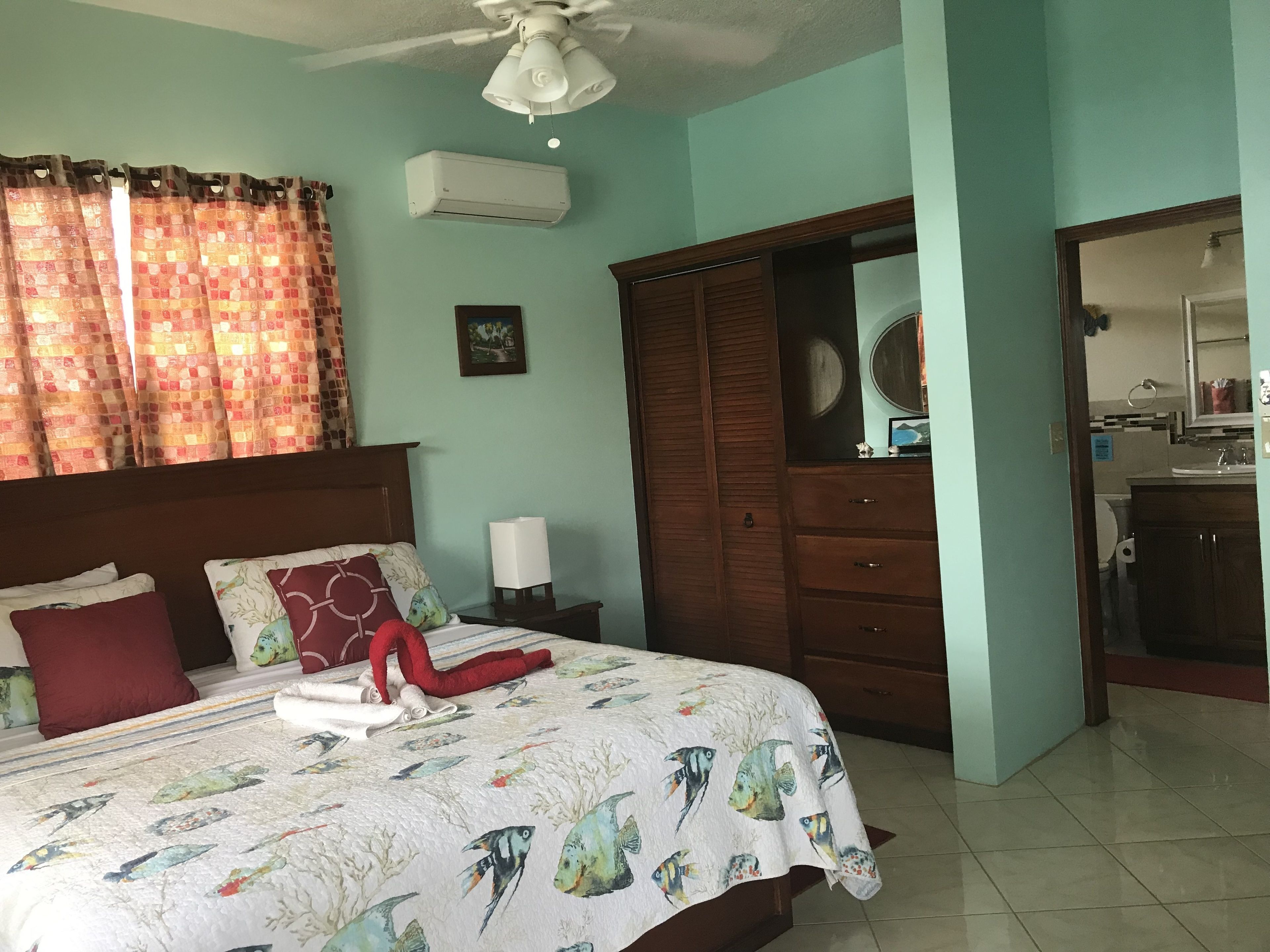 Une chambre à coucher avec un lit, des rideaux, un ventilateur de plafond, un climatiseur, une armoire et une salle de bain visible à travers une porte ouverte.