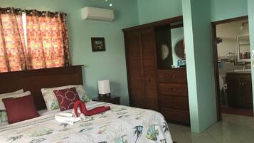 Une chambre à coucher avec un lit, des rideaux, un ventilateur de plafond, un climatiseur, une armoire et une salle de bain visible à travers une porte ouverte.