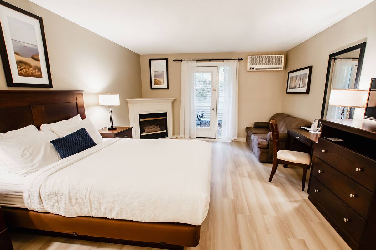 A well-lit bedroom with a large bed, a dresser, a chair, a fireplace, and a view through a glass door.