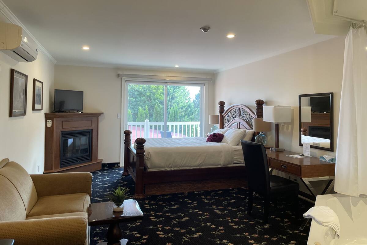 A hotel room with a bed, a desk, a chair, a TV, a fireplace, and a balcony with a view of trees.