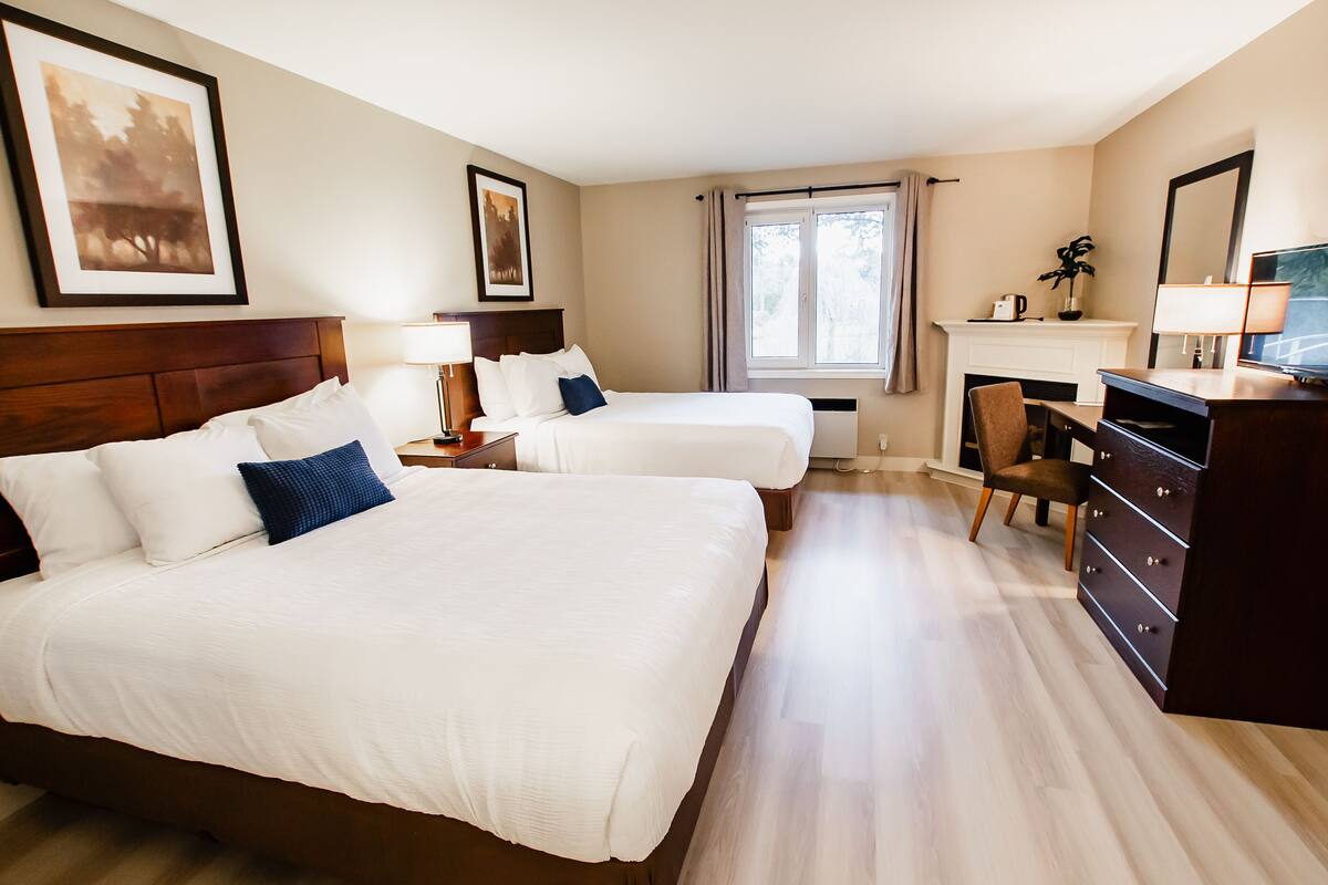 A hotel room with two beds, a desk, a chair, a dresser, a fireplace, and framed pictures on the walls.