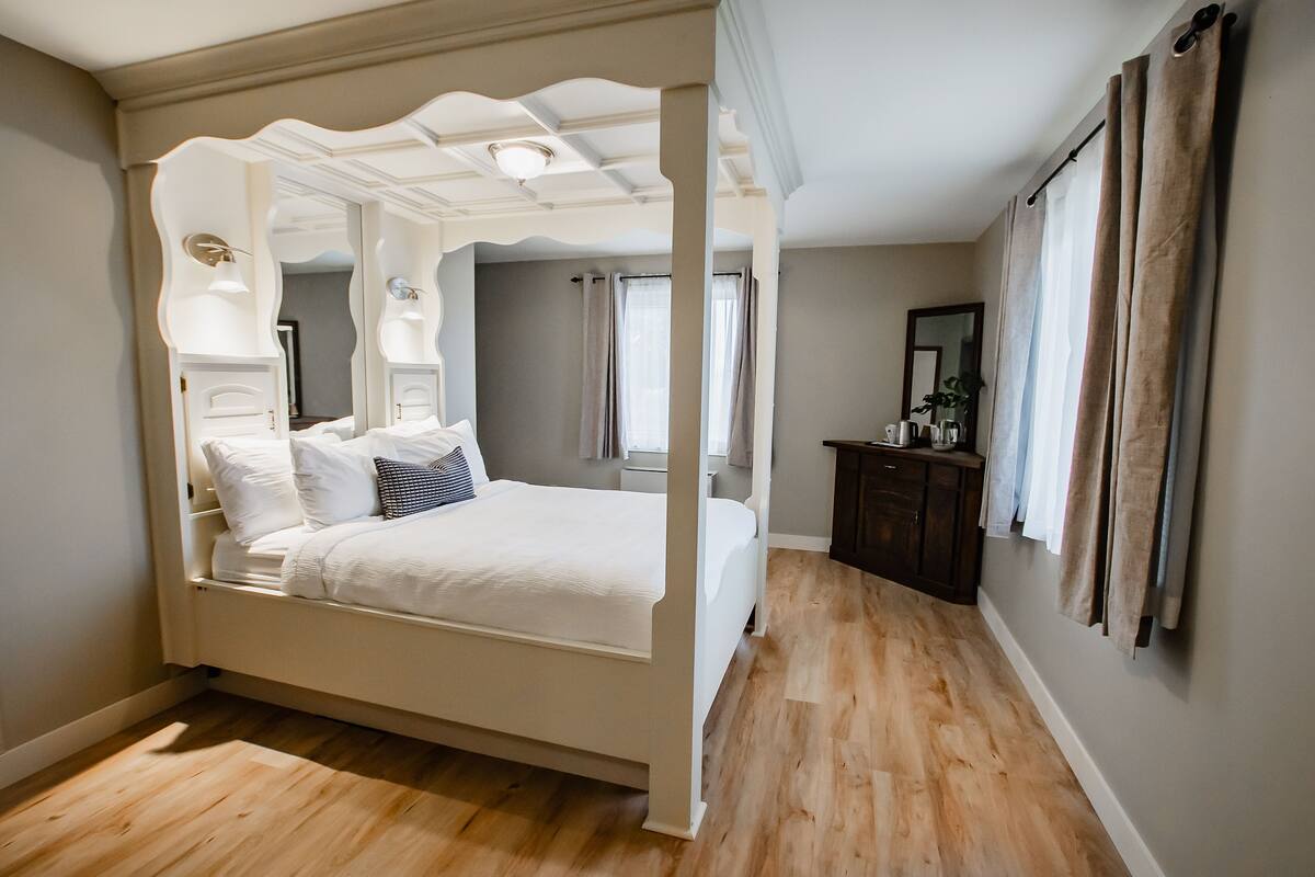 A four-poster bed in a bedroom with a wooden floor, a dresser, and a mirror on the wall.