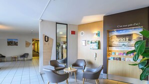 A hotel lobby with a reception desk, seating area with chairs and a small table, a large mirror, and a wall with framed pictures and a poster. - B&B HOTEL Orange (Orange)
