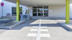 A modern building entrance with a covered waiting area, a wheelchair-accessible path, and a no parking sign. - B&B HOTEL Orange (Orange)