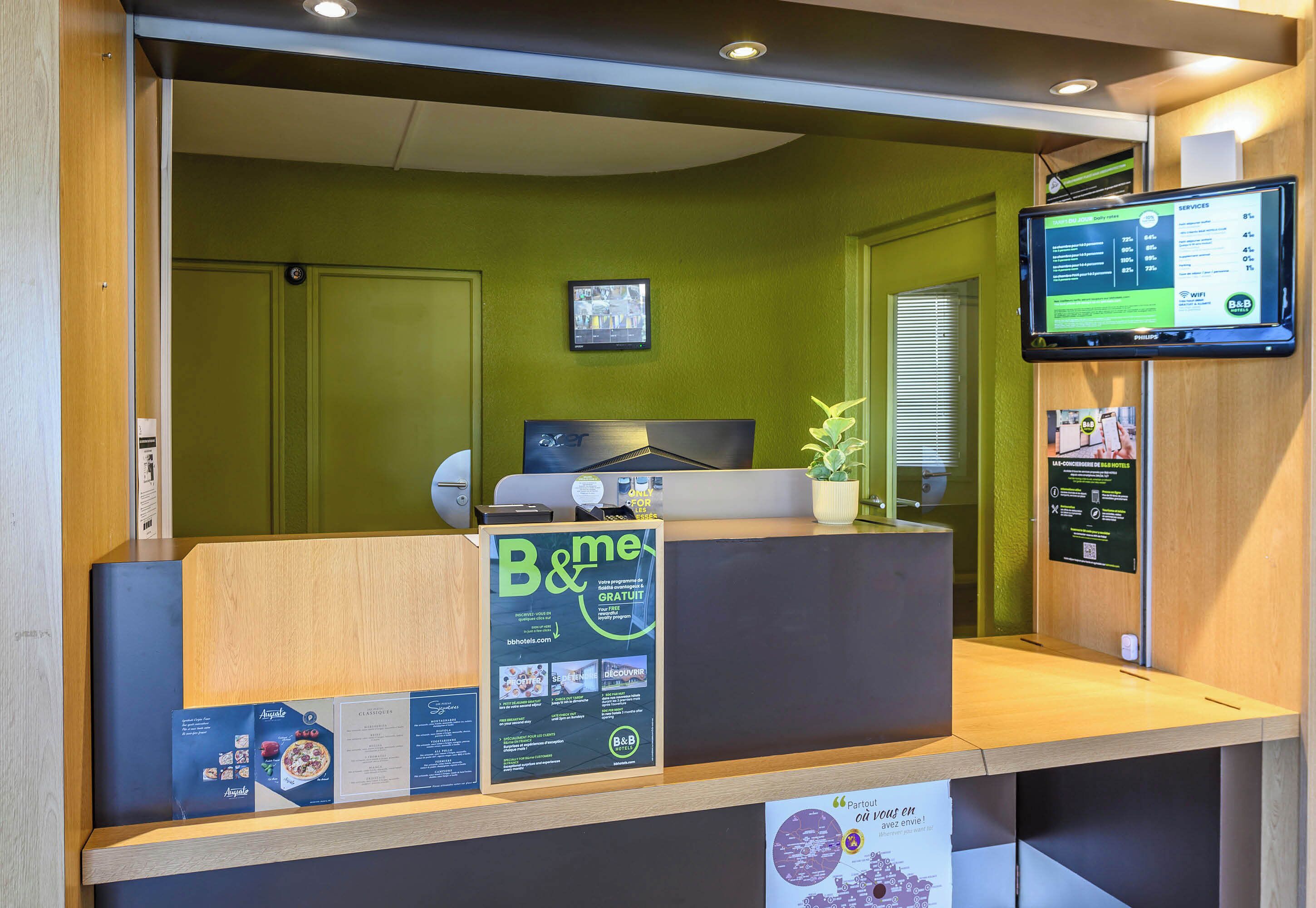 Un comptoir d’accueil avec un écran affichant des informations, un panneau « B&Me » et une petite plante.