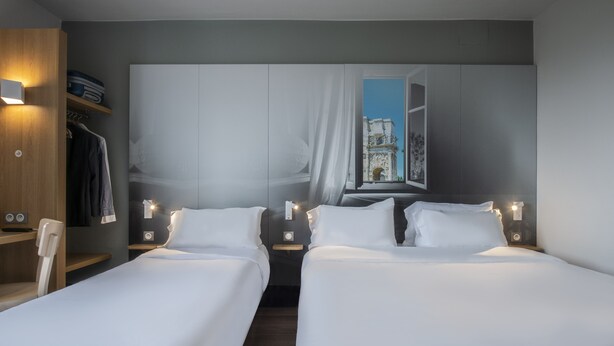 A hotel room with two single beds, a wardrobe, a desk, and a window with a view of a historic building. - B&B HOTEL Orange (Orange)