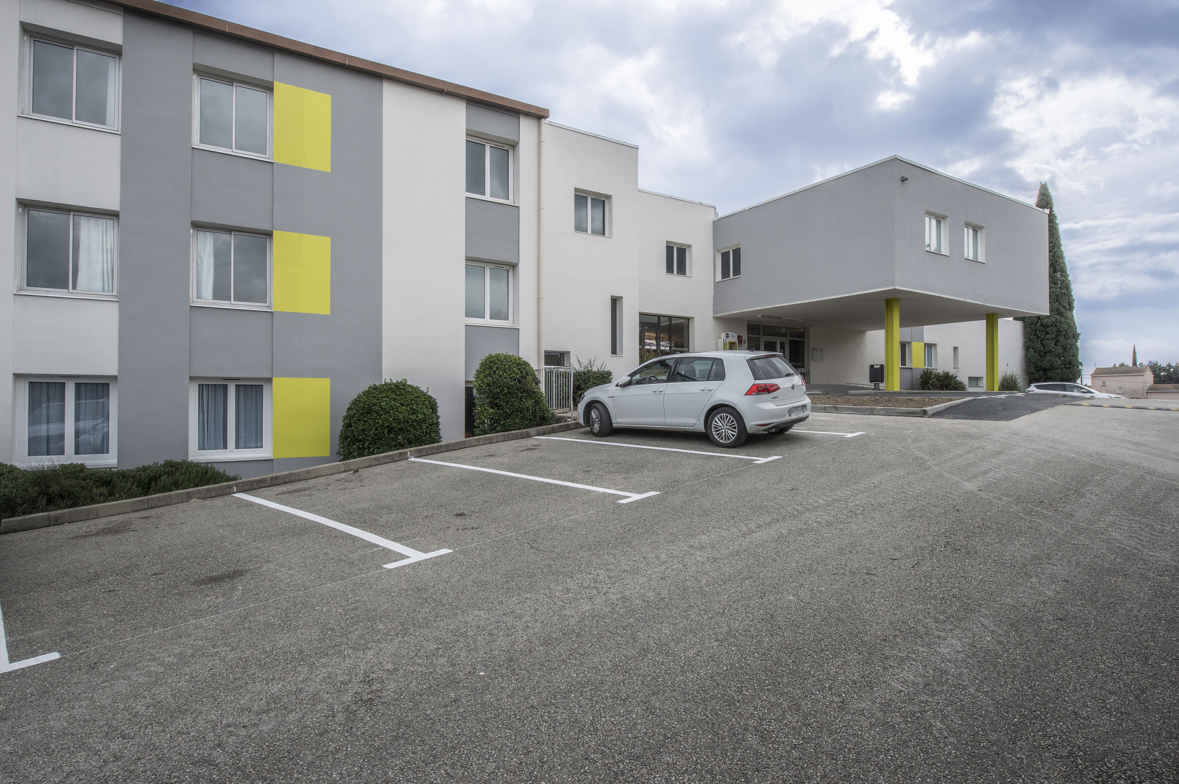 Un bâtiment moderne avec un parking, une voiture blanche et, au fond, un bâtiment jaune.