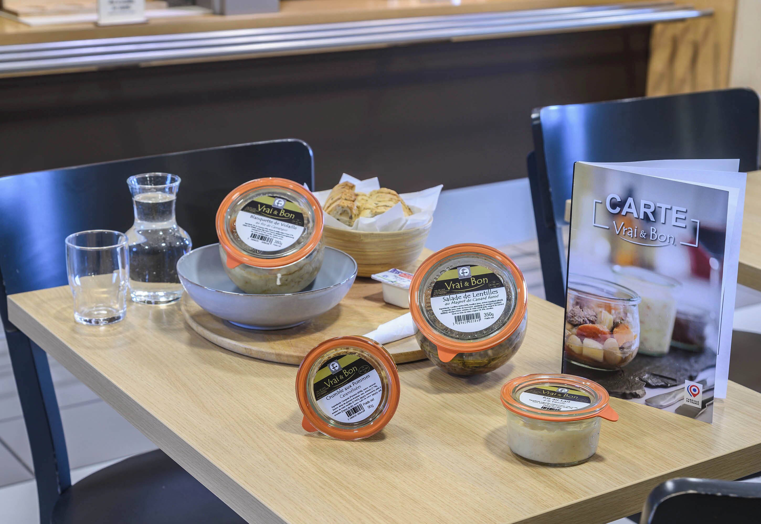 Una mesa con frascos de alimentos en conserva, un cuenco de pan y un cartel con el menú.